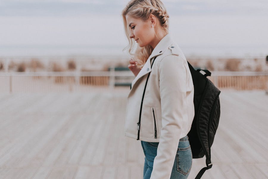 Woman with blonde braids and backpack walking on a wooden boardwalk with blurred seaside background.