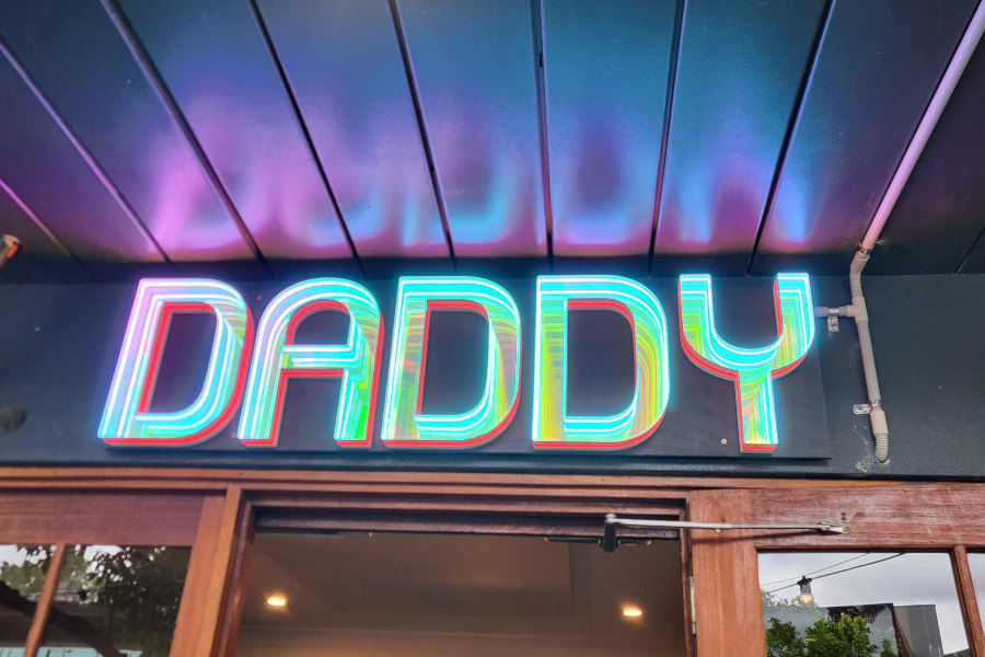 Exterior view of Come to Daddy bar showing a colourful neon sign reading “Daddy” above the entrance with wooden doors and ceiling panels.