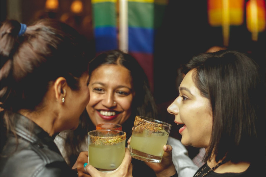 Three women smiling and toasting with cocktails at Sidecar bar, with rainbow flag in background.