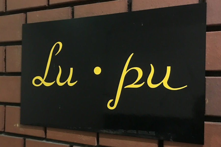 Lu Pu Bar logo displayed in yellow script on a black signboard attached to a textured brick wall.