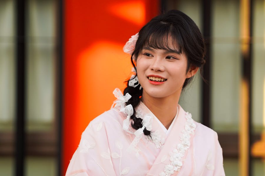 ピンクの花柄の着物を着た若いトランスMtF。編み込みの髪にヘアクリップをつけ、屋外で微笑んでいる。