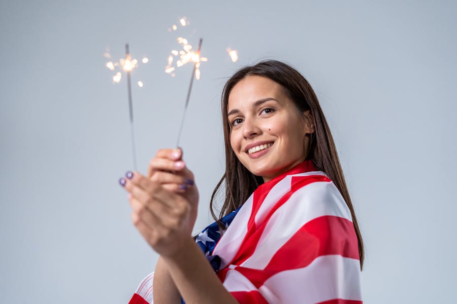 Smiling woman wrapped in U.S. flag holding sparklers, celebrating with a light blue background.