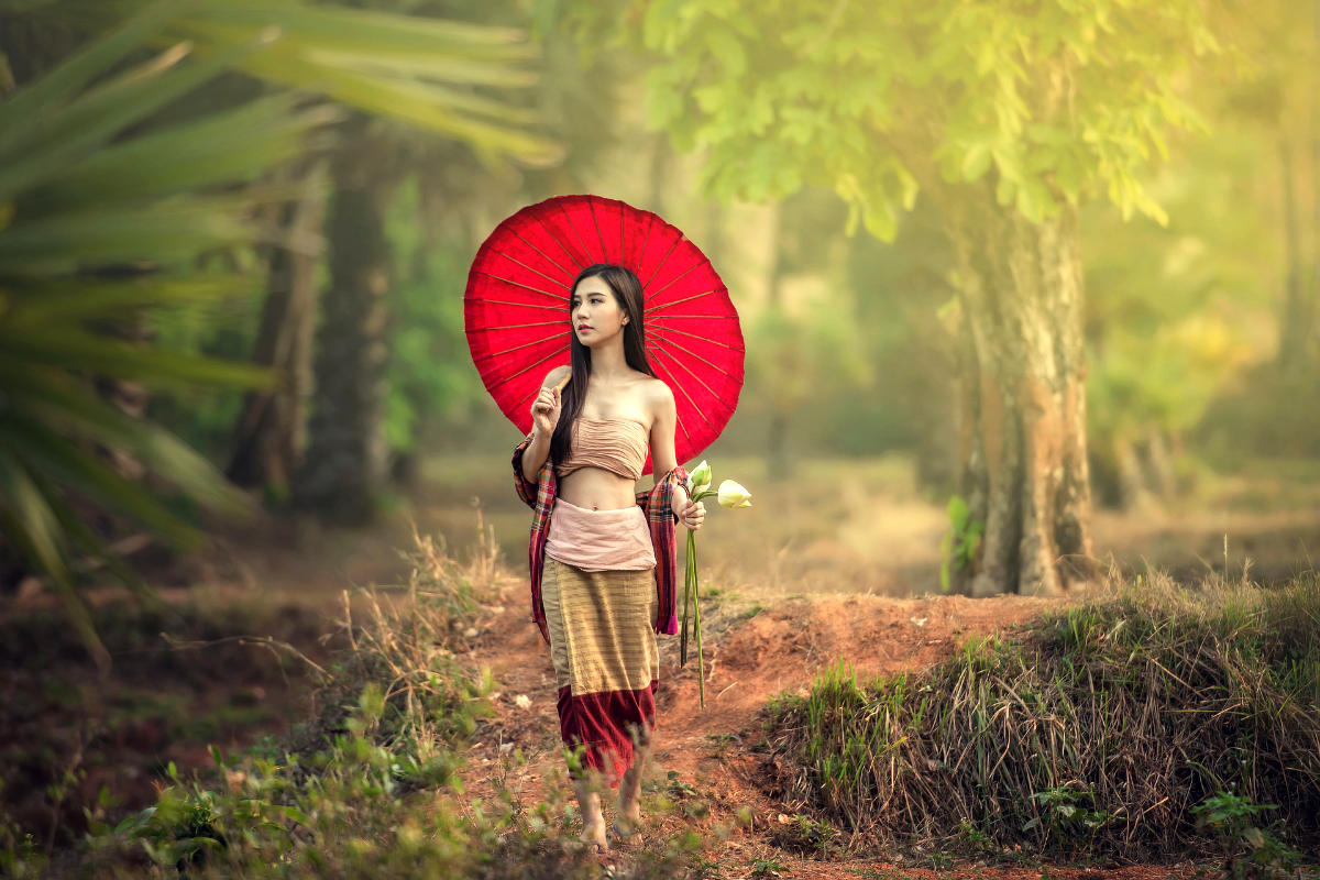 A trans woman in traditional Thai dress stands on a forest path, holding a lotus flower and red parasol in soft sunlight.