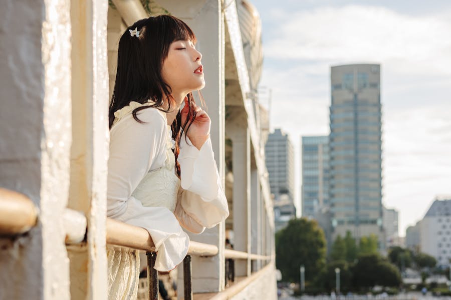 白い服を着たトランスMtFが、目を閉じてバルコニーの手すりにもたれている。背後には、柔らかな光に包まれた街の建物が広がっている。